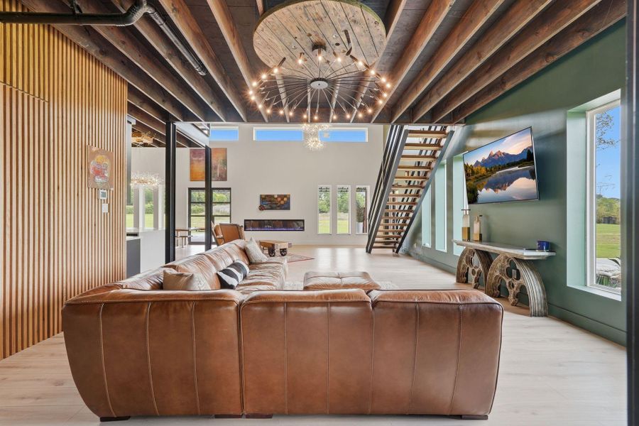 This modern living space features a high ceiling with exposed beams and a unique chandelier, a cozy brown leather sectional, and an open staircase. Large windows provide ample natural light and offer scenic views, enhancing the room's inviting atmosphere.