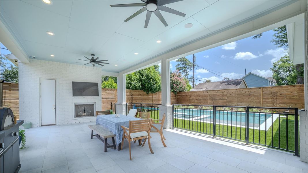 This photo showcases a spacious, covered outdoor patio featuring a ceiling fan, fireplace, and mounted TV. There’s a dining area with a table and chairs, overlooking a fenced backyard with a pool, perfect for entertaining and relaxation.
