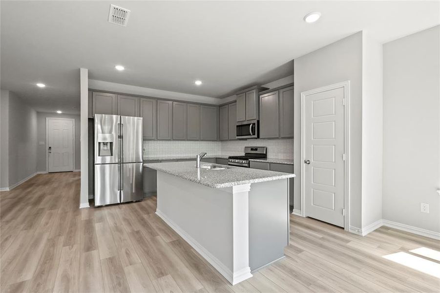 Kitchen with light stone counters, an island with sink, light hardwood / wood-style floors, appliances with stainless steel finishes, and decorative backsplash