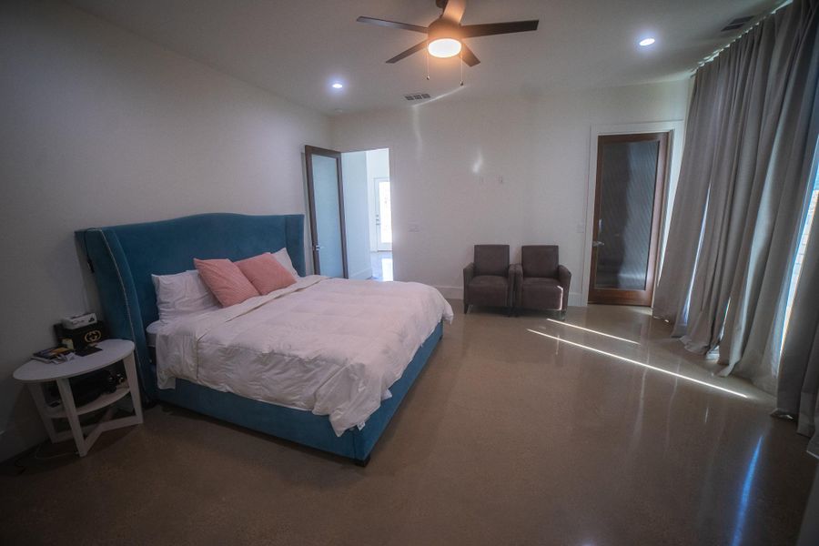 Spacious bedroom with polished floors, a large bed with a blue headboard, and soft lighting. The room features modern ceiling fan, two armchairs, and floor-to-ceiling curtains, offering a cozy and inviting atmosphere.
