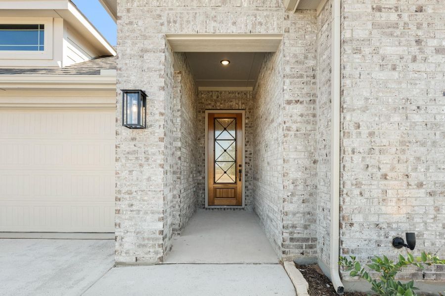 Covered front entry with wood door, glass panels, and light brick exterior walls beside a garage