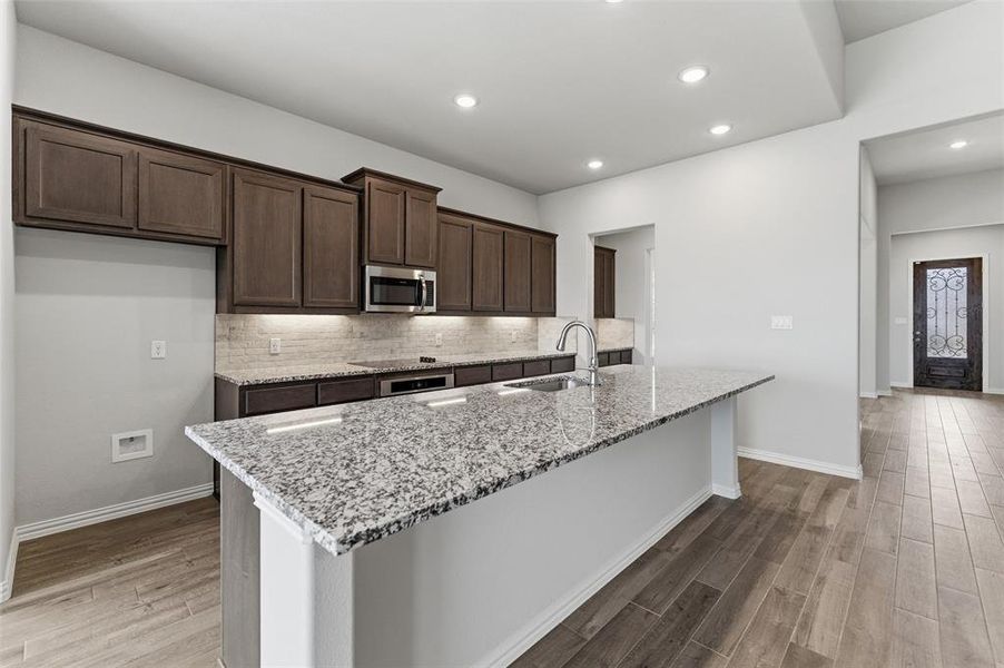 Kitchen with dark wood finish cabinets, light stone countertops, an island with sink, light wood finished floors, and tasteful backsplash