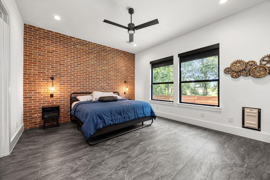 This spacious Primary bedroom features a modern industrial design with a striking brick accent wall, large windows for natural light, sleek tile flooring, and contemporary lighting fixtures. This spacious Primary bedroom features a modern industrial design with a striking brick accent wall, large windows for natural light, sleek tile flooring, and contemporary lighting fixtures.