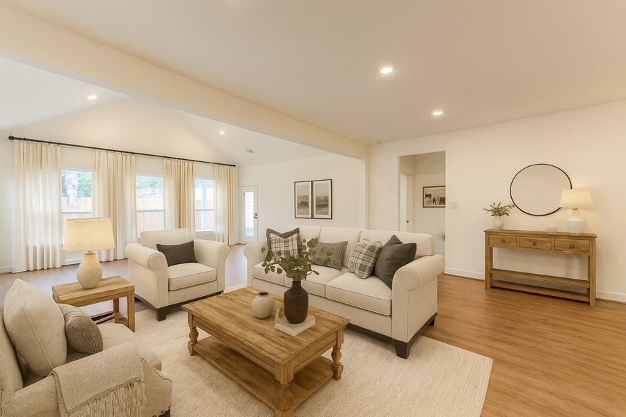 Bright and spacious living room with vaulted ceilings, neutral tones, and elegant wood flooring. Features comfortable seating, a wooden coffee table, and abundant natural light from large windows with sheer curtains.