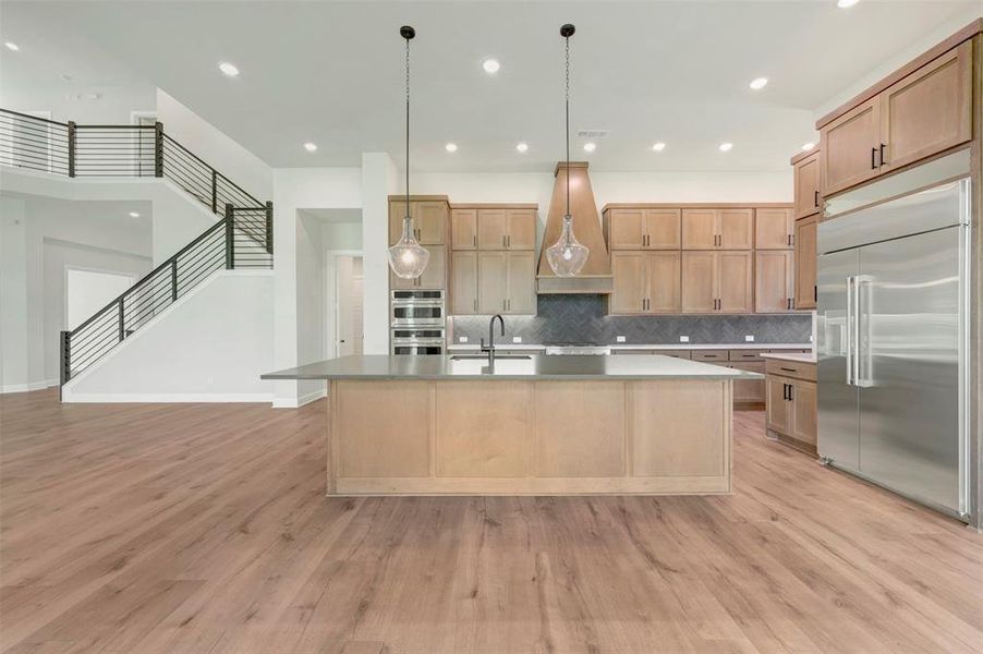 Kitchen with stainless steel appliances, a center island with sink, tasteful backsplash, light wood-style flooring, and decorative light fixtures