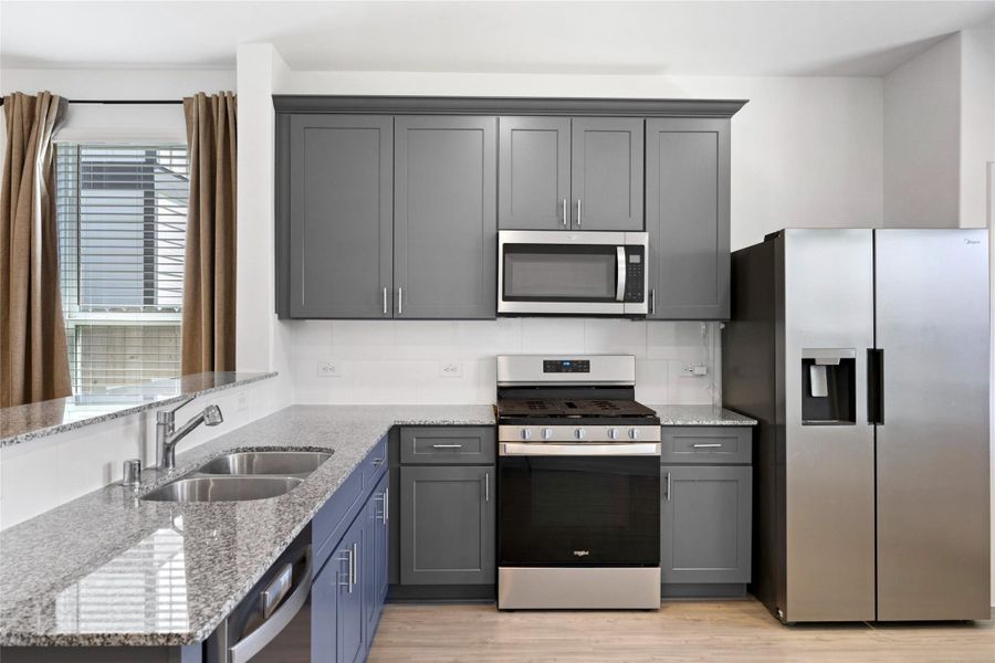 modern kitchen features sleek gray cabinetry, stainless steel appliances, a granite countertop, and a double sink. The space is brightened by a window with curtains, offering a stylish and functional cooking area.