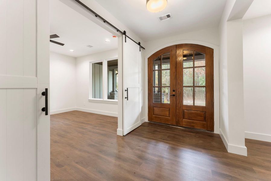 This photo showcases a bright entryway with elegant double wooden doors and glass panels, leading to the study and dining room with plank floors and a large window, creating a welcoming space.
