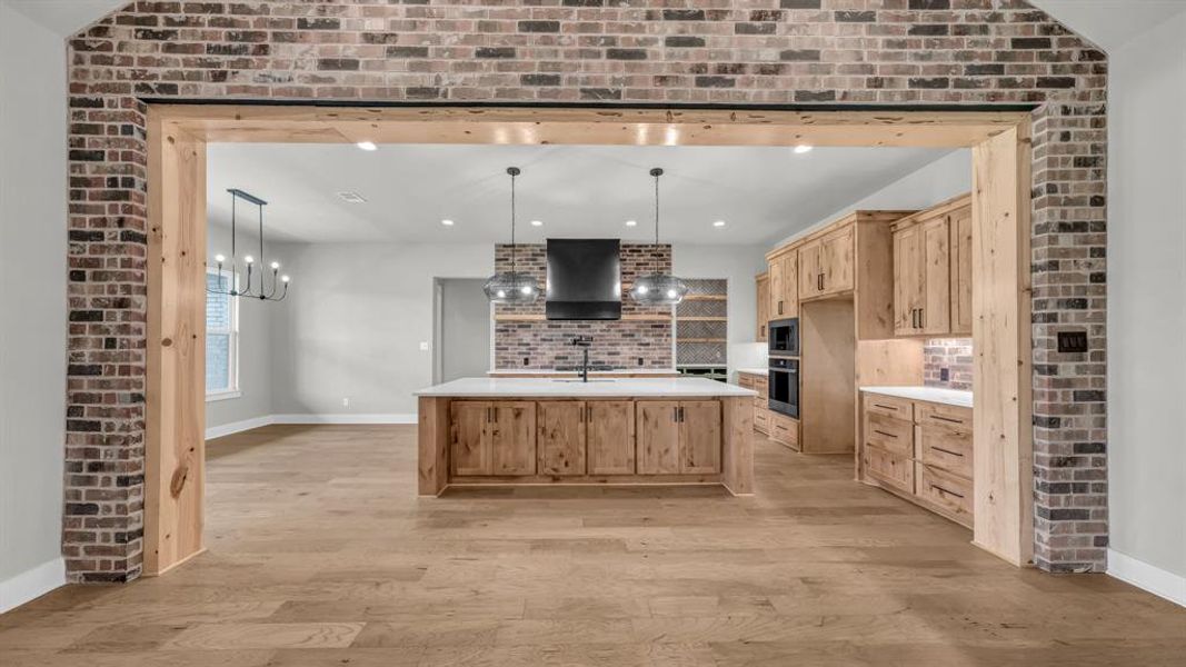 Brick archway with wood beam framing leading to a kitchen featuring a central island with white countertop, wood cabinetry, and stainless steel appliances