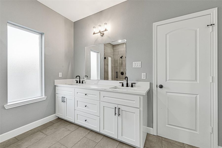 This bathroom features a modern double vanity with sleek black fixtures, a large mirror, and ample storage. The neutral gray walls and tiled floor create a clean, contemporary look, while a frosted window allows natural light to filter in.
