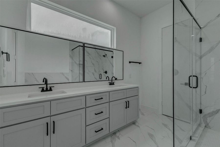 The primary bathroom features a long dual-sink vanity with soft gray cabinetry, generous drawer and cabinet storage, matte black fixtures, and a wide black-framed mirror. A glass-enclosed shower with marble-look tile and black hardware creates a bright, spa-inspired focal point