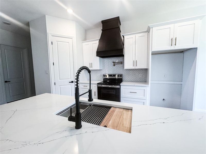 Kitchen featuring stainless steel range with electric cooktop, custom range hood, backsplash, white cabinetry, and light stone countertops Kitchen featuring stainless steel range with electric cooktop, custom range hood, backsplash, white cabinetry, and light stone countertops