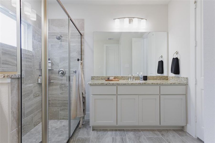 Bathroom featuring a spacious walk-in shower with glass enclosure, light gray tile flooring, a white vanity with ample storage, a granite-style countertop, and a large mirror with contemporary lighting