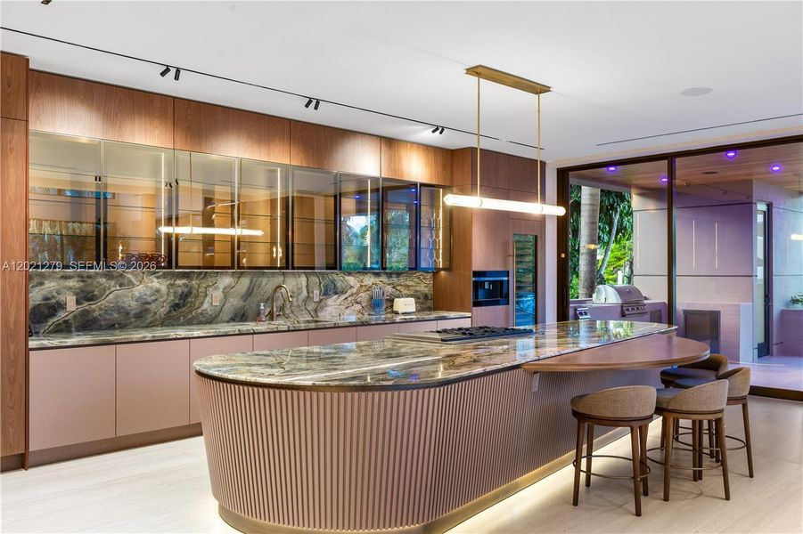 Designer kitchen with curved stone island, under-island LED lighting, fluted panel base, smoked glass display cabinets, walnut upper cabinetry, dramatic stone backsplash, brass pendant fixture, and sliding glass door opening to covered outdoor kitchen and pool area.
