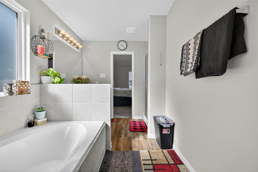 This photo showcases a modern bathroom with a large soaking tub, tiled walls, and a window providing natural light. It features stylish decor, including plants and a towel rack, and has an adjoining door leading to a bedroom. The space is bright and welcoming, offering a cozy yet contemporary feel.