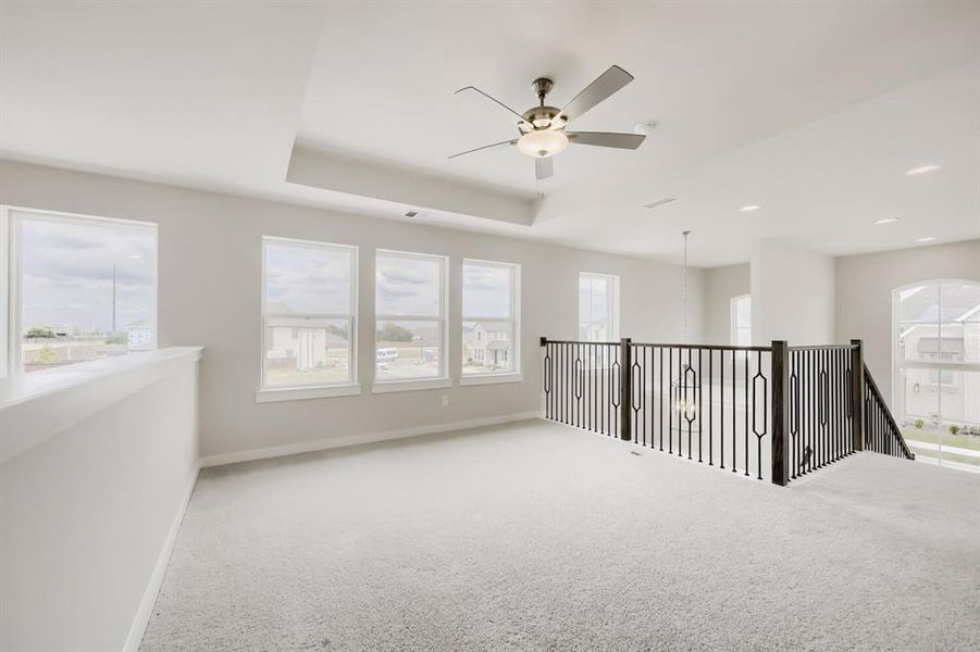 Spacious upper-level area featuring light-toned carpeting, multiple large windows, a tray ceiling, and a ceiling fan with integrated lighting