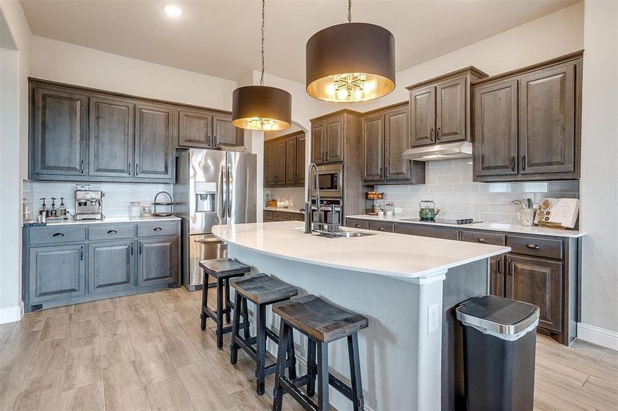 Kitchen with backsplash, a kitchen island with sink, hanging light fixtures, dark brown cabinets, and stainless steel appliances