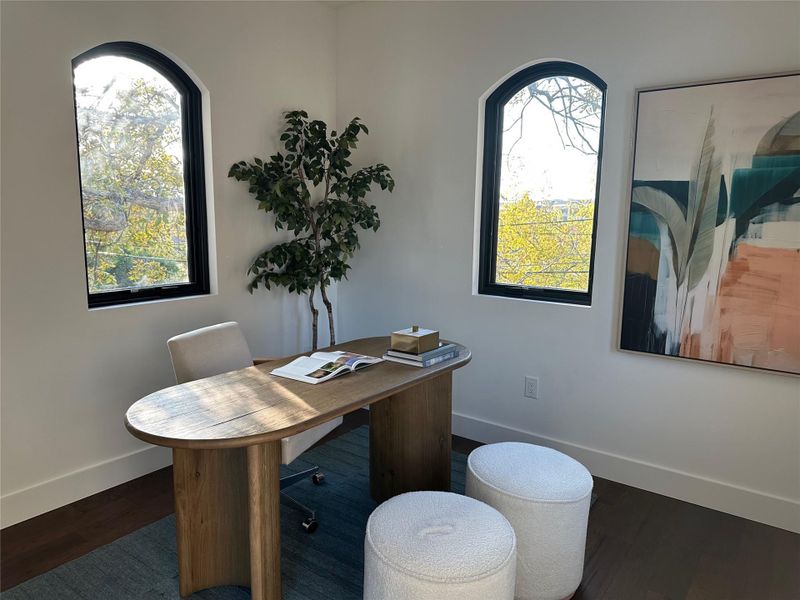 Room featuring two arched windows with black frames, white walls, dark wood-finish flooring, and a contemporary light wood desk