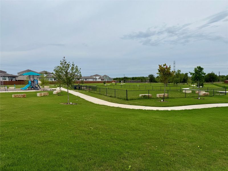 Expansive green space featuring a concrete pathway, a fenced pet area, and a playground with slides and climbing structures