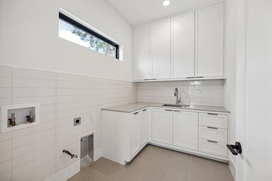 This upstairs utility room has a clean, minimalistic design with bright white cabinetry and countertops. The space includes a built-in sink with ample counter space for laundry-related tasks.