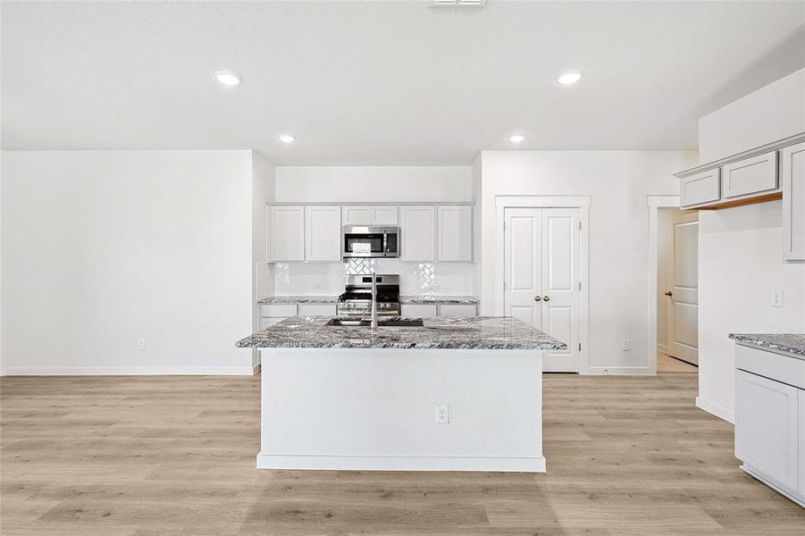 Kitchen with light wood-type flooring, appliances with stainless steel finishes, decorative backsplash, light stone counters, and an island with sink