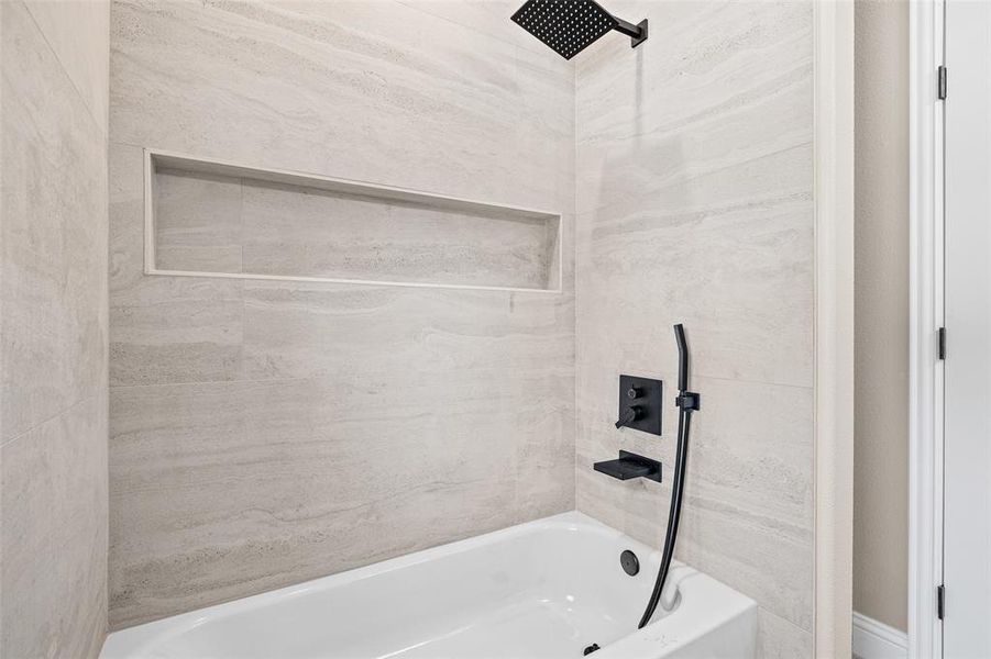 Bathtub with light-toned tiled surround, matte black rainfall showerhead, handheld shower fixture, and recessed shower niche
