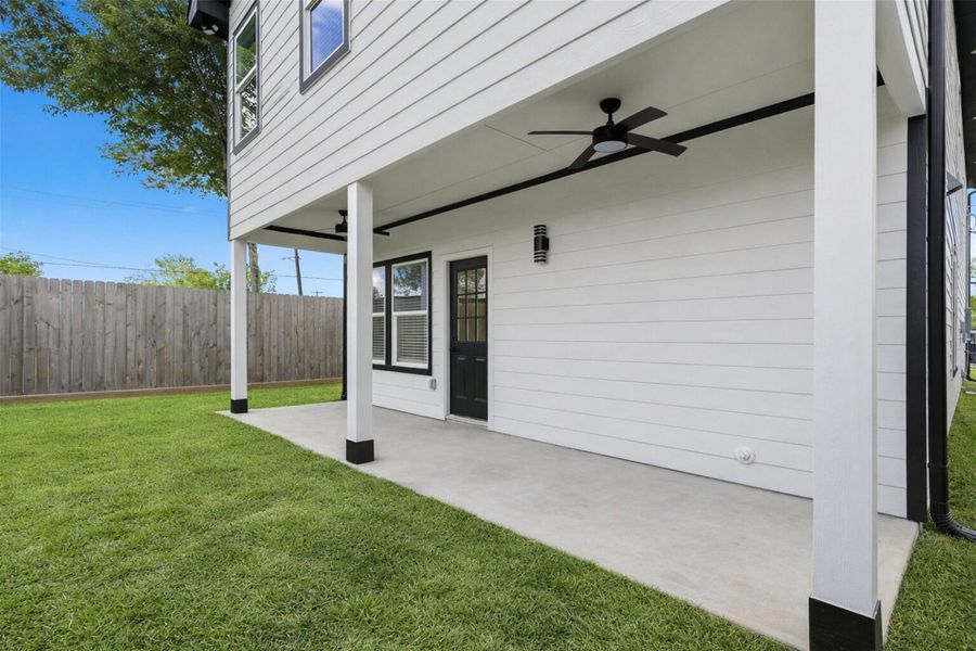 Covered back patio with ceiling fan overlooking a lush green backyard enclosed by a privacy fence.