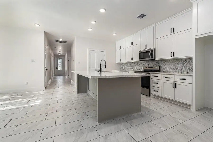 This modern kitchen features sleek white cabinetry, a stylish tile backsplash, and stainless steel appliances. The spacious island provides ample counter space, while the open layout and light tile flooring enhance the bright, airy atmosphere.