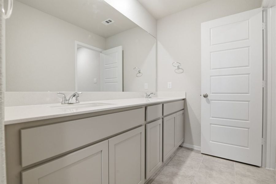 Image of a bathroom with light grey cabinets, a white jack and jill vanity, a large mirror and tile flooring