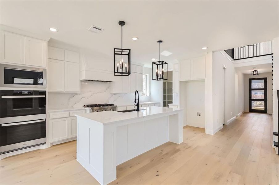 Kitchen with tasteful backsplash, white cabinetry, a kitchen island with sink, light wood-type flooring, and decorative light fixtures