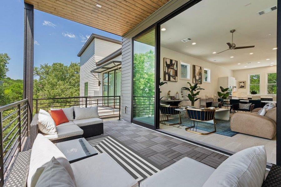 Seamless indoor-outdoor living space featuring oversized sliding glass doors opening to a spacious covered balcony with skyline and treetop views.
