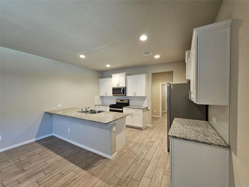 Kitchen with white cabinetry, open floor plan, stainless steel appliances, recess lights, granite counters, backsplash and wood looking tiled floors.