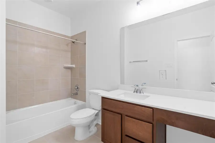 Secondary bath features tile flooring, bath/shower combo with tile surround, stained cabinetry with beautiful light countertops, large mirror with sleek fixtures.