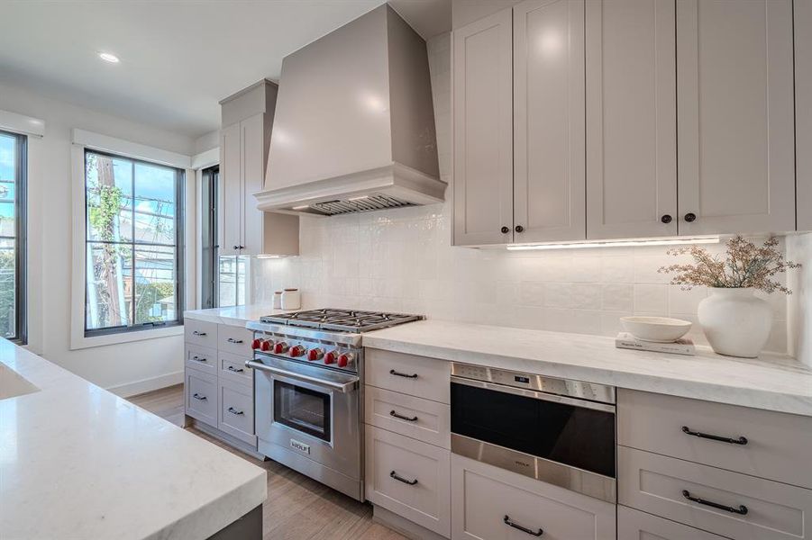 Kitchen featuring premium range hood, appliances with stainless steel finishes, backsplash, light stone counters, and light wood finished floors