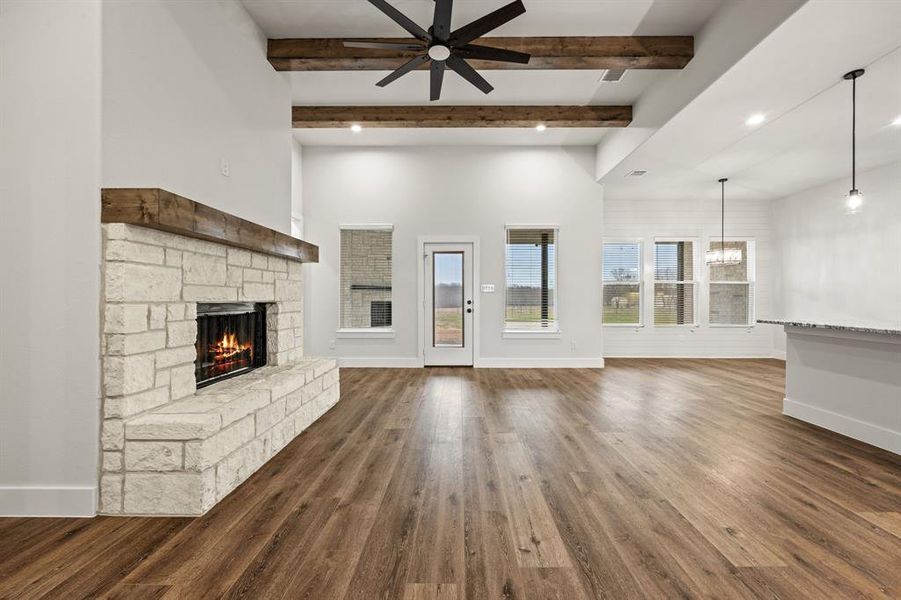 Unfurnished living room featuring baseboards, beamed ceiling, dark wood-type flooring, a stone fireplace, and ceiling fan with notable chandelier