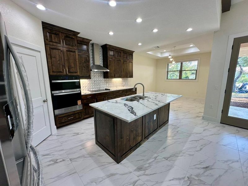 Kitchen with dark brown cabinetry, decorative backsplash, fridge with ice dispenser, a raised ceiling, and a kitchen island with sink