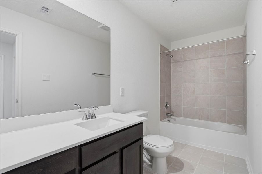 The secondary bathroom offers tile flooring, a bath/shower combination with a stylish tile surround, rich stained wood cabinetry with added knee space and elegant light countertops, a mirror, and sleek fixtures with modern finishes.