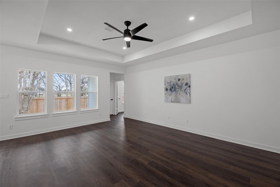 Family room features dark hardwood floors, a tray ceiling with recessed lighting, and a modern ceiling fan. Large windows provide natural light and a view of the fenced backyard, creating a bright and inviting space. Family room features dark hardwood floors, a tray ceiling with recessed lighting, and a modern ceiling fan. Large windows provide natural light and a view of the fenced backyard, creating a bright and inviting space.