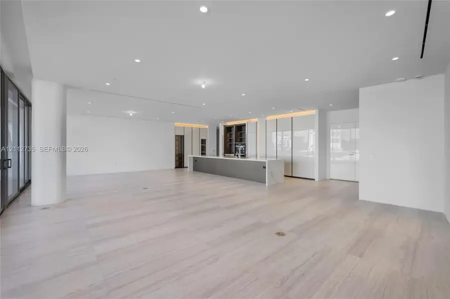 Open-concept space featuring light-toned wood-finish flooring, recessed ceiling lighting, floor-to-ceiling glass sliding doors, and a kitchen island with a stone-finish countertop and integrated cabinetry