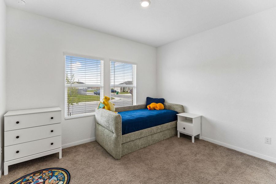 A well-lit bedroom features plush carpeting and two large windows with horizontal blinds.