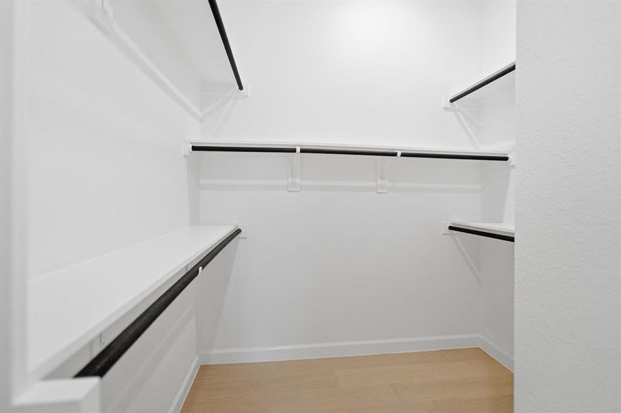 Walk-in closet featuring light wood-finish flooring, white walls, and a multi-tiered shelving system with integrated black hanging rods Walk-in closet featuring light wood-finish flooring, white walls, and a multi-tiered shelving system with integrated black hanging rods