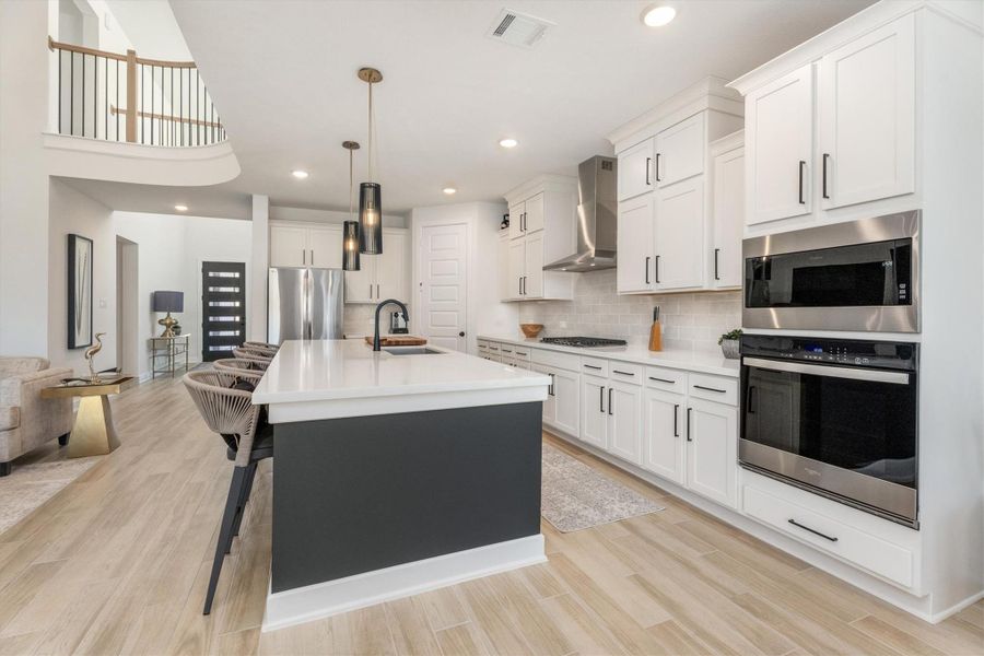 This modern kitchen features sleek white cabinetry and stainless steel appliances, including a double oven. The large island with seating offers ample workspace and is complemented by stylish pendant lighting. The open layout seamlessly connects to the living area, creating a spacious and welcoming environment.