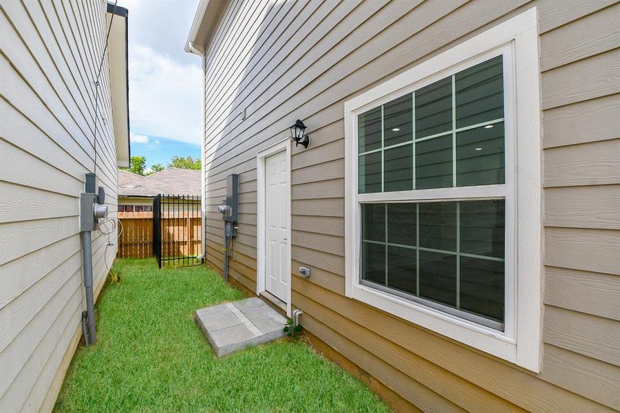 This photo shows a well-maintained side yard with a small grassy area, a concrete stepping stone path, a side entrance door, and a large window, all enclosed by a wooden privacy fence. Ideal for outdoor utility space or a private garden area.