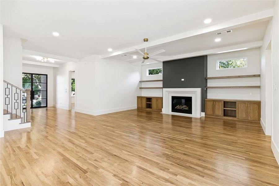 Spacious room featuring wood-finish flooring, a contemporary fireplace with a dark accent wall, built-in cabinetry, floating shelves, and a ceiling fan with integrated lighting Spacious room featuring wood-finish flooring, a contemporary fireplace with a dark accent wall, built-in cabinetry, floating shelves, and a ceiling fan with integrated lighting