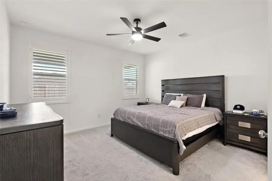 This bedroom features a modern, minimalist design with a dark wood bed frame and matching nightstands. It has neutral-colored walls, ample natural light from two windows, and a ceiling fan for comfort.