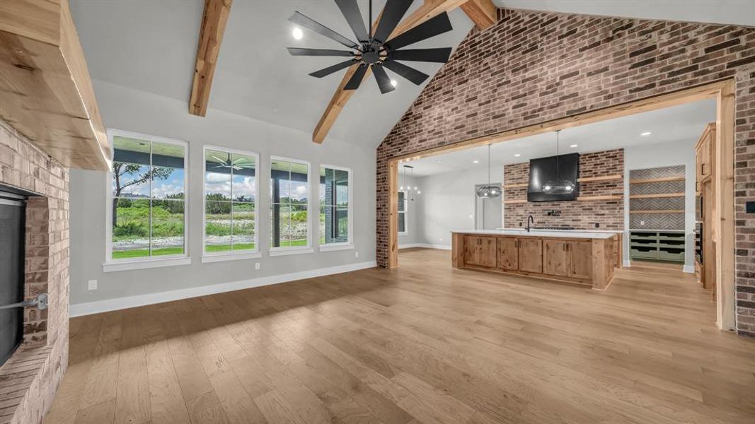 Vaulted ceiling with exposed wood beams, brick accent wall, wood-finish flooring, multiple windows, and a large ceiling fan