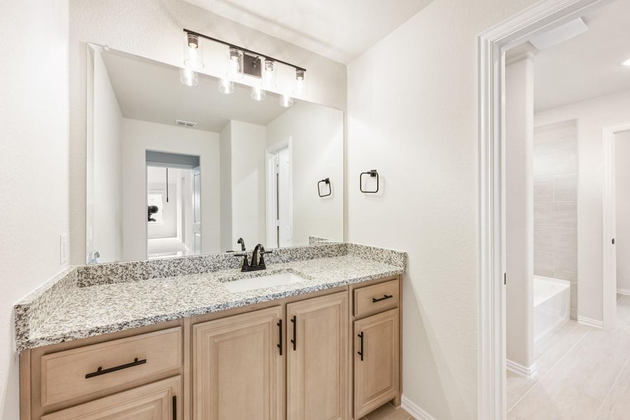 Bathroom vanity with granite countertop, light wood cabinets, large mirror, and soaking tub visible in background Bathroom vanity with granite countertop, light wood cabinets, large mirror, and soaking tub visible in background