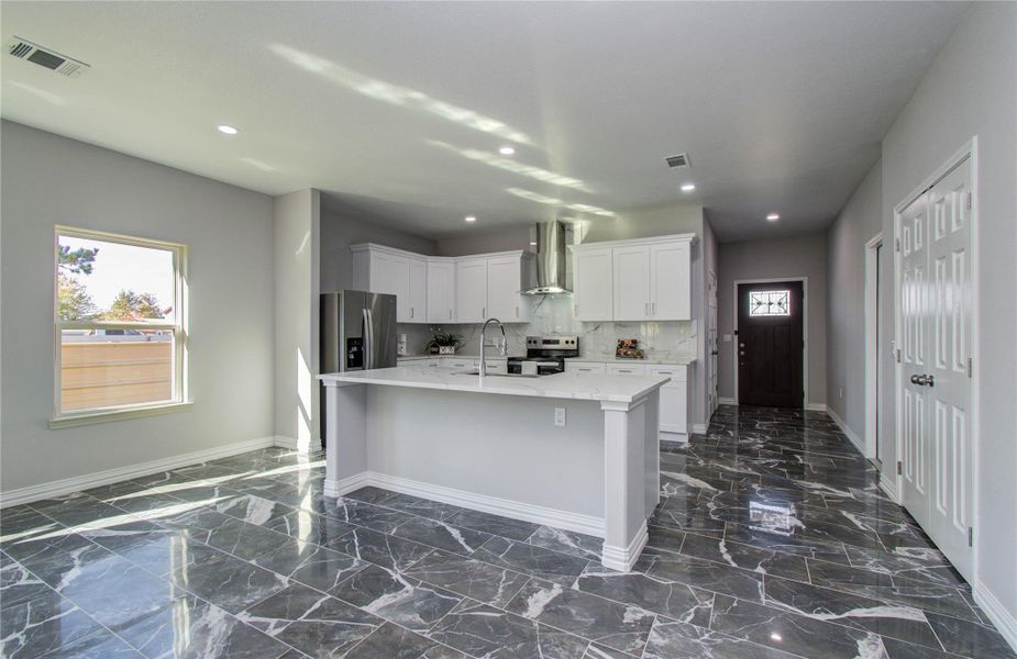 Modern kitchen with sleek white cabinetry and stainless steel appliances. Features a central island with marble countertops and glossy gray tile flooring. Ample natural light streams through a large window, enhancing the open, inviting space.