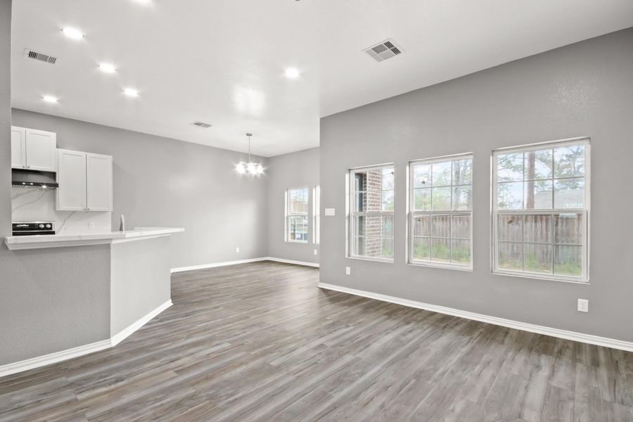 This bright and open living space features sleek wood flooring, neutral gray walls, and ample natural light from large windows. The adjacent kitchen area includes modern lighting and a bar counter, creating a welcoming atmosphere for entertaining or relaxation.