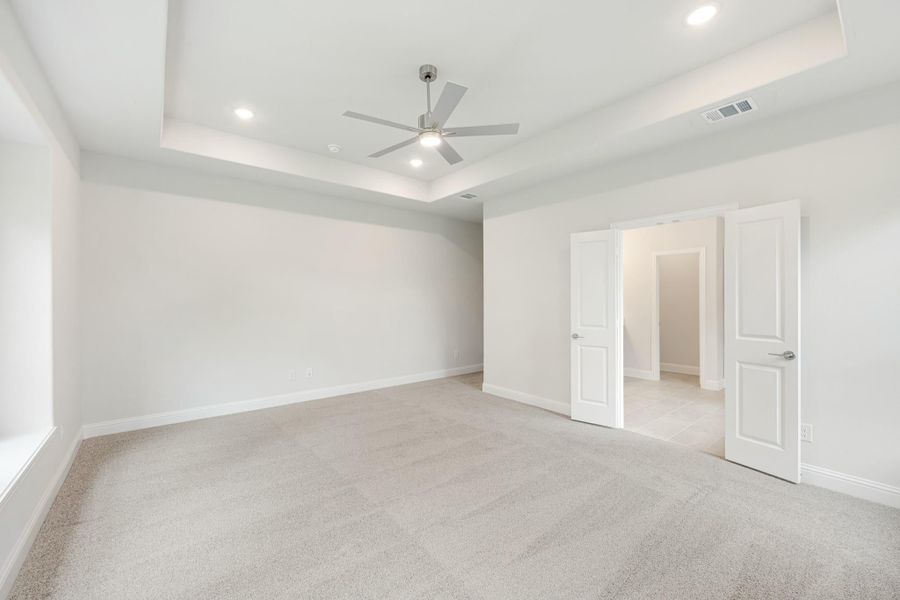Empty bedroom with tray ceiling, ceiling fan, carpet flooring, and two white doors leading to bathroom and hallway Empty bedroom with tray ceiling, ceiling fan, carpet flooring, and two white doors leading to bathroom and hallway