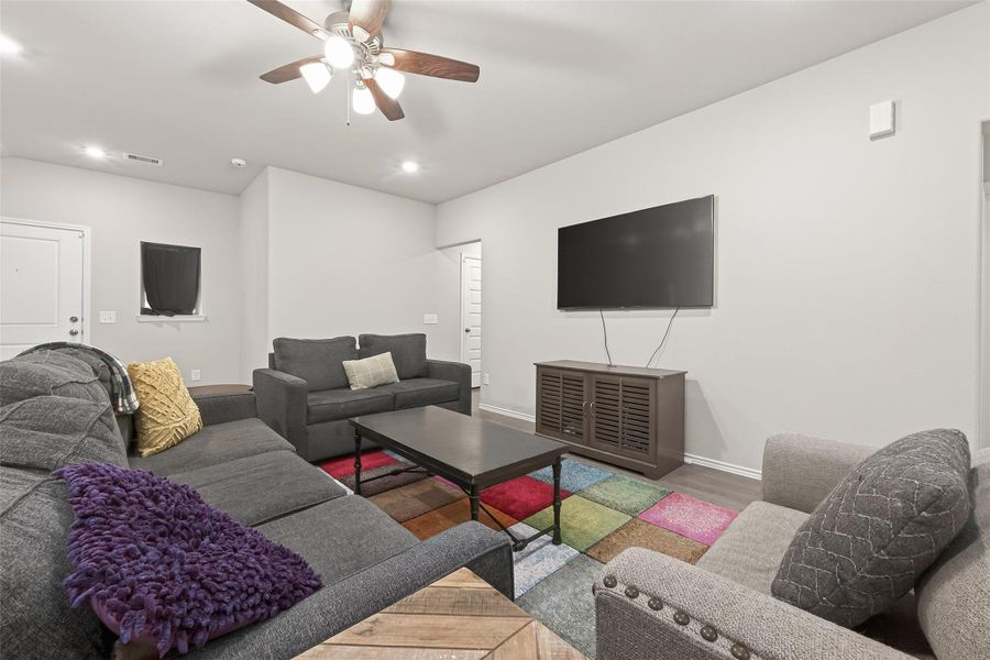 This living room features a cozy seating arrangement with a sectional sofa and armchair, a wall-mounted TV, and a colorful area rug. The space is well-lit with ceiling lights and a fan, creating a welcoming atmosphere.
